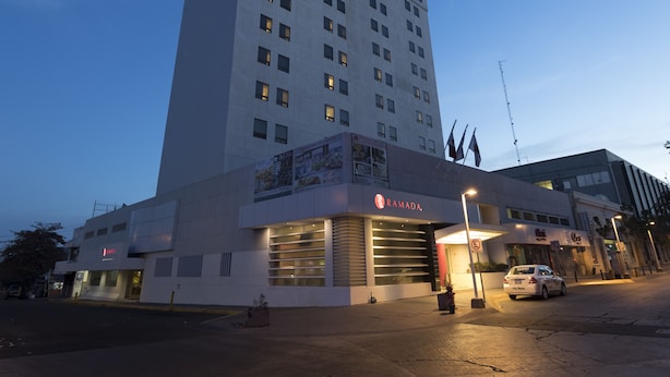 Un hotel moderno con un llamativo letrero de 'RAMADA', un estacionamiento amplio y un edificio alto al fondo.