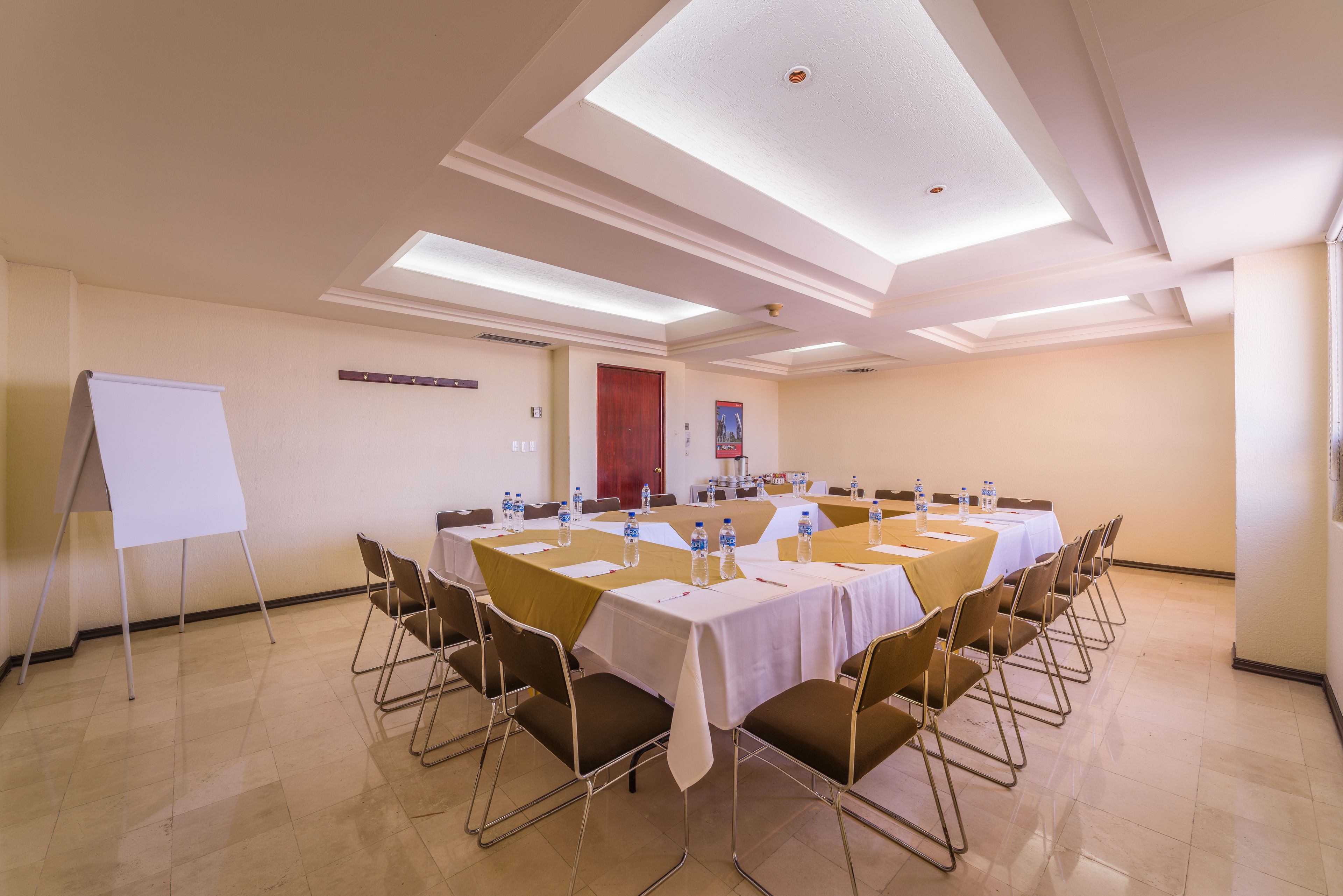 Un salón de conferencias con una pizarra, mesas con sillas y botellas de agua.
