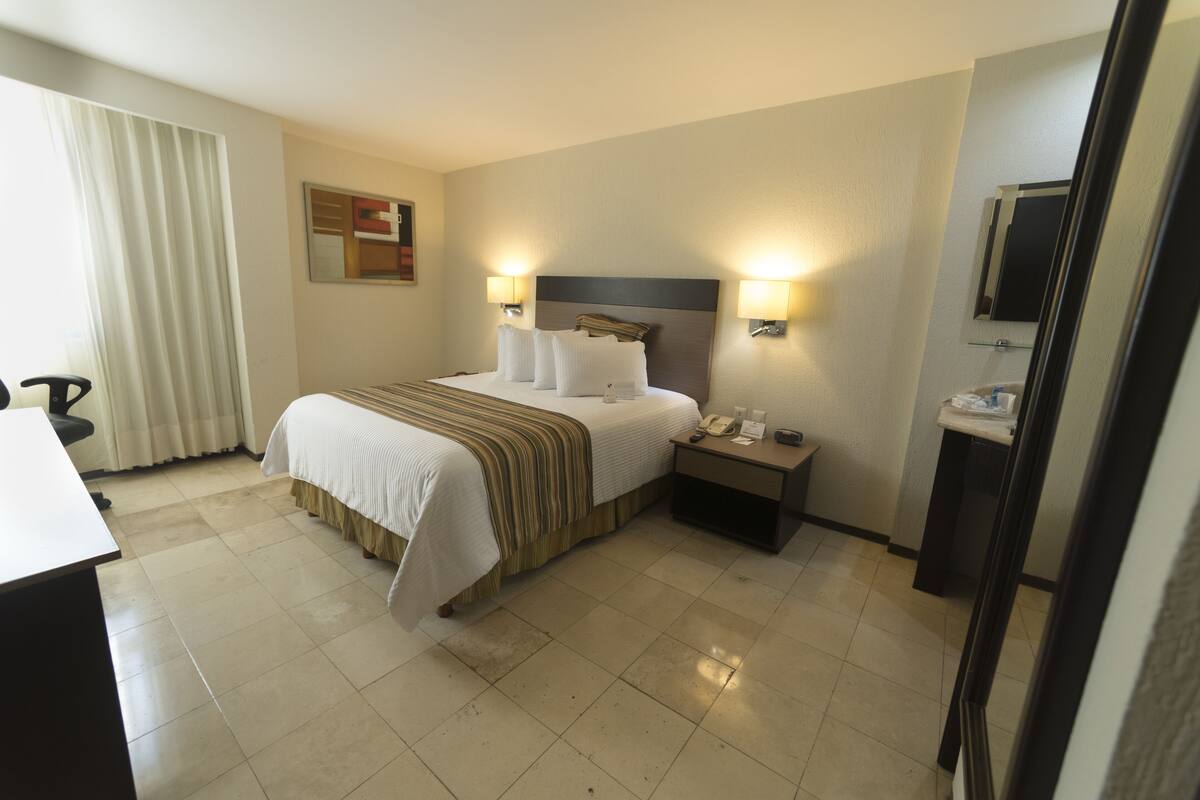 Una habitación de hotel con cama, lámparas de noche, mesita de noche, escritorio y espejo.