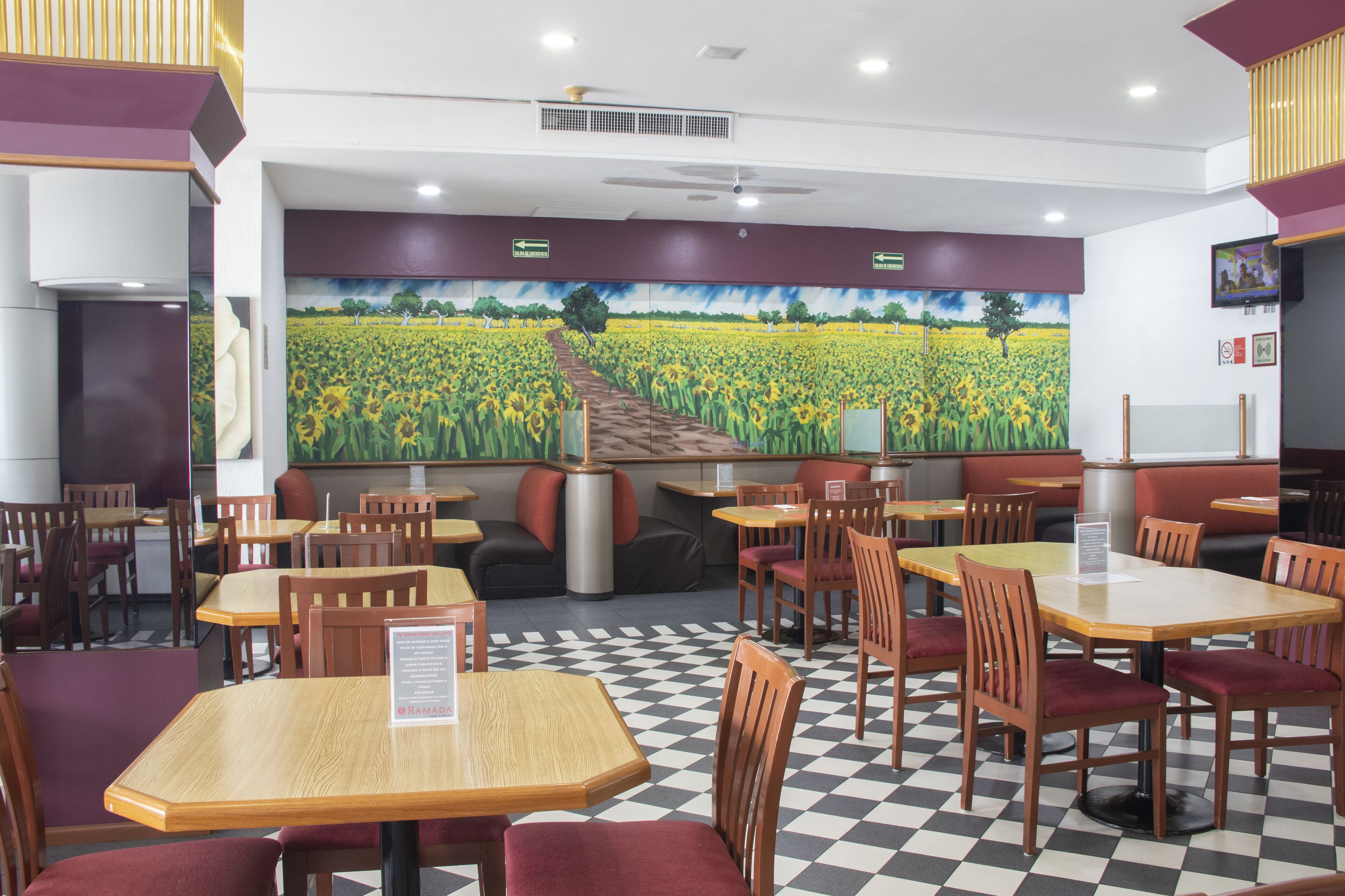 A restaurant with checkered flooring, wooden tables and chairs, and a mural of a field on the back wall.