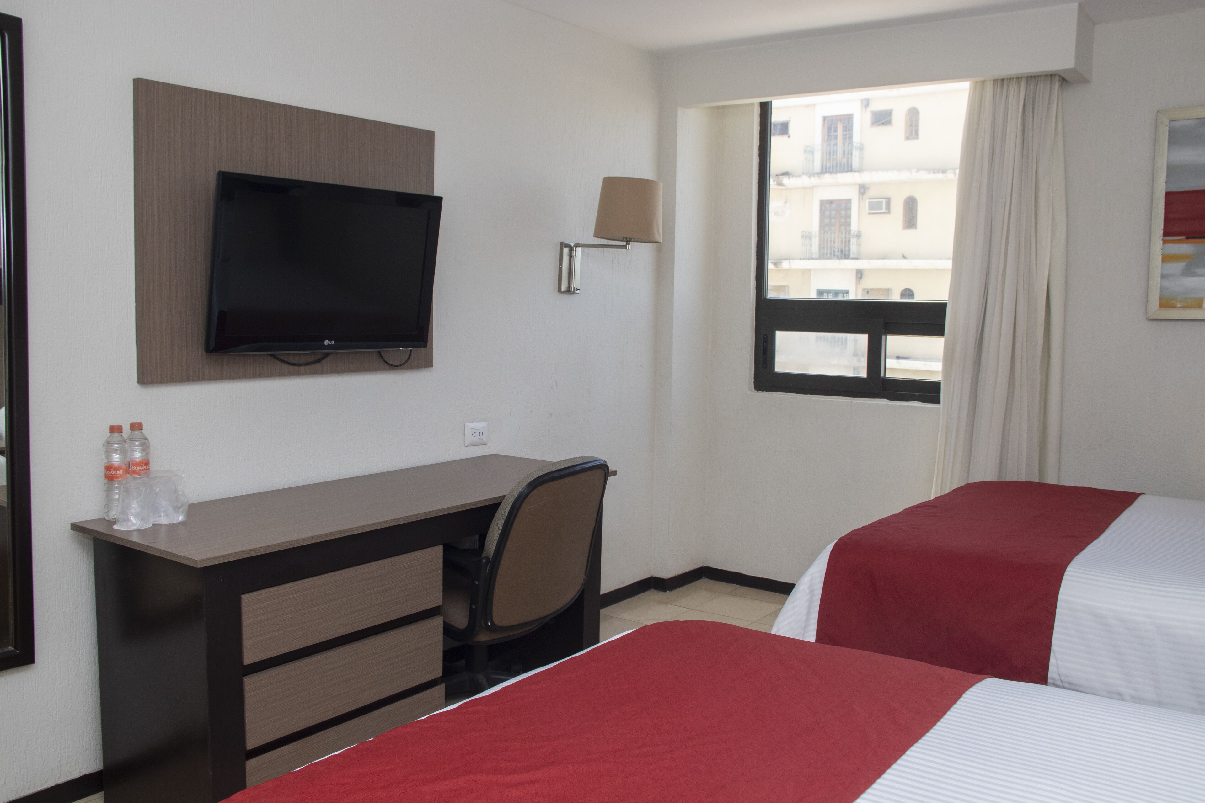 Una habitación de hotel con cama, escritorio, silla, televisor y ventana con cortinas.