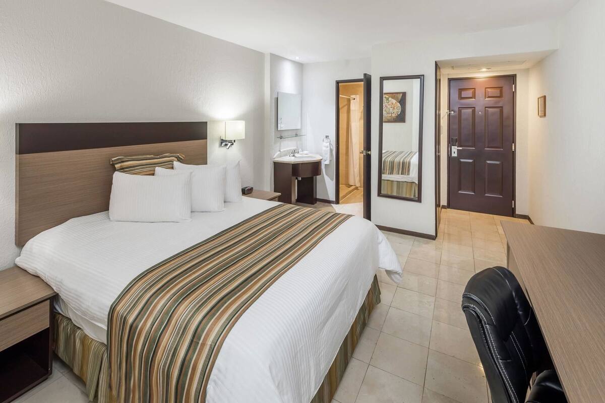 Habitación de hotel con cama, mesita de noche, escritorio, silla y baño con lavamanos y espejo.