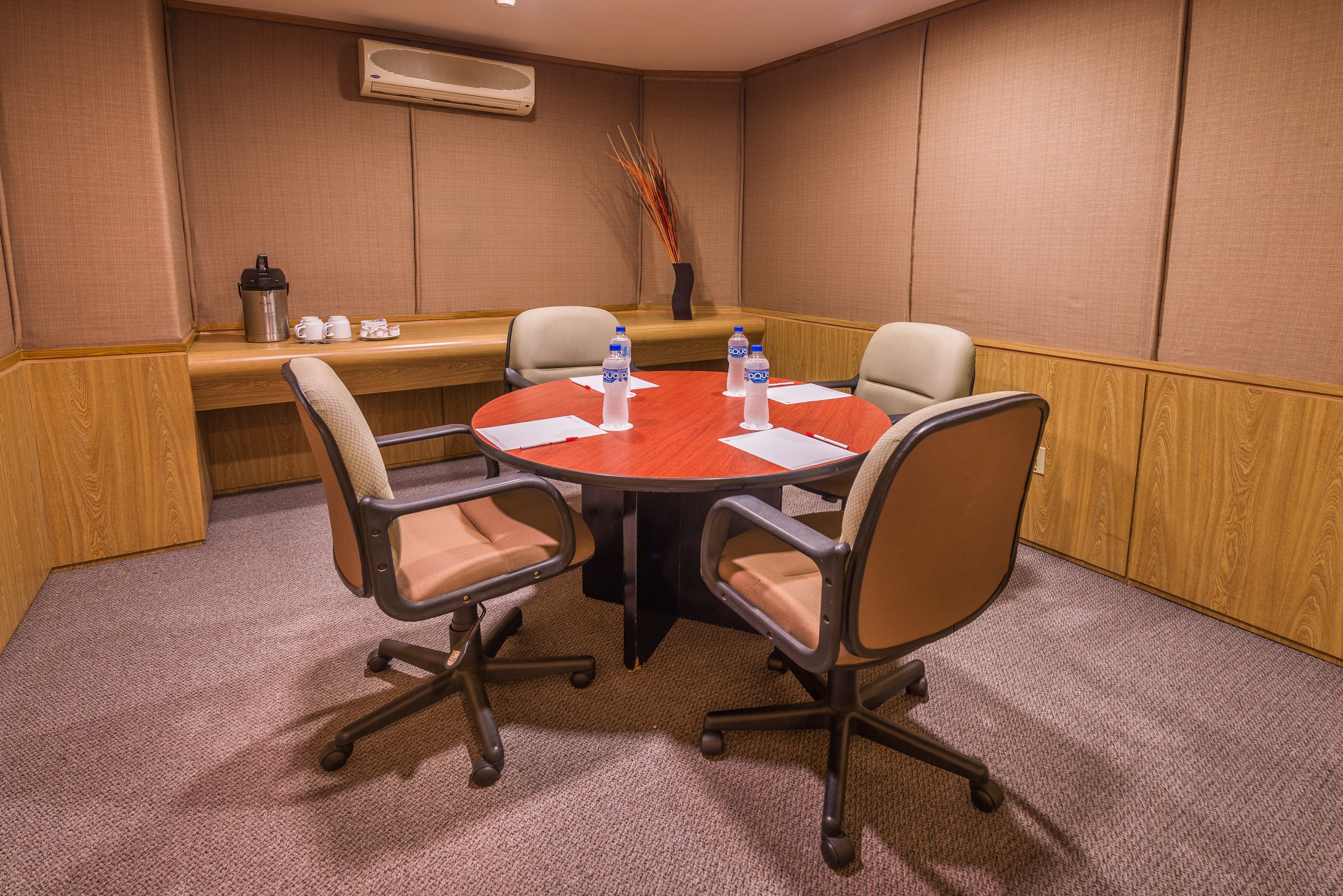 A conference room with a round table, four chairs, a coffee station, and a vase with decorative sticks.
