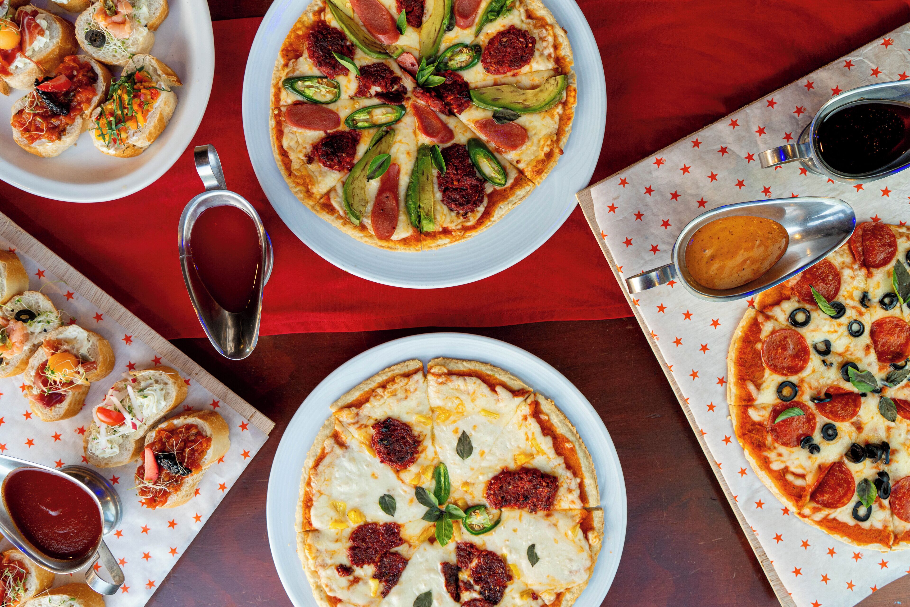A variety of pizzas and appetizers on a table with a red tablecloth.