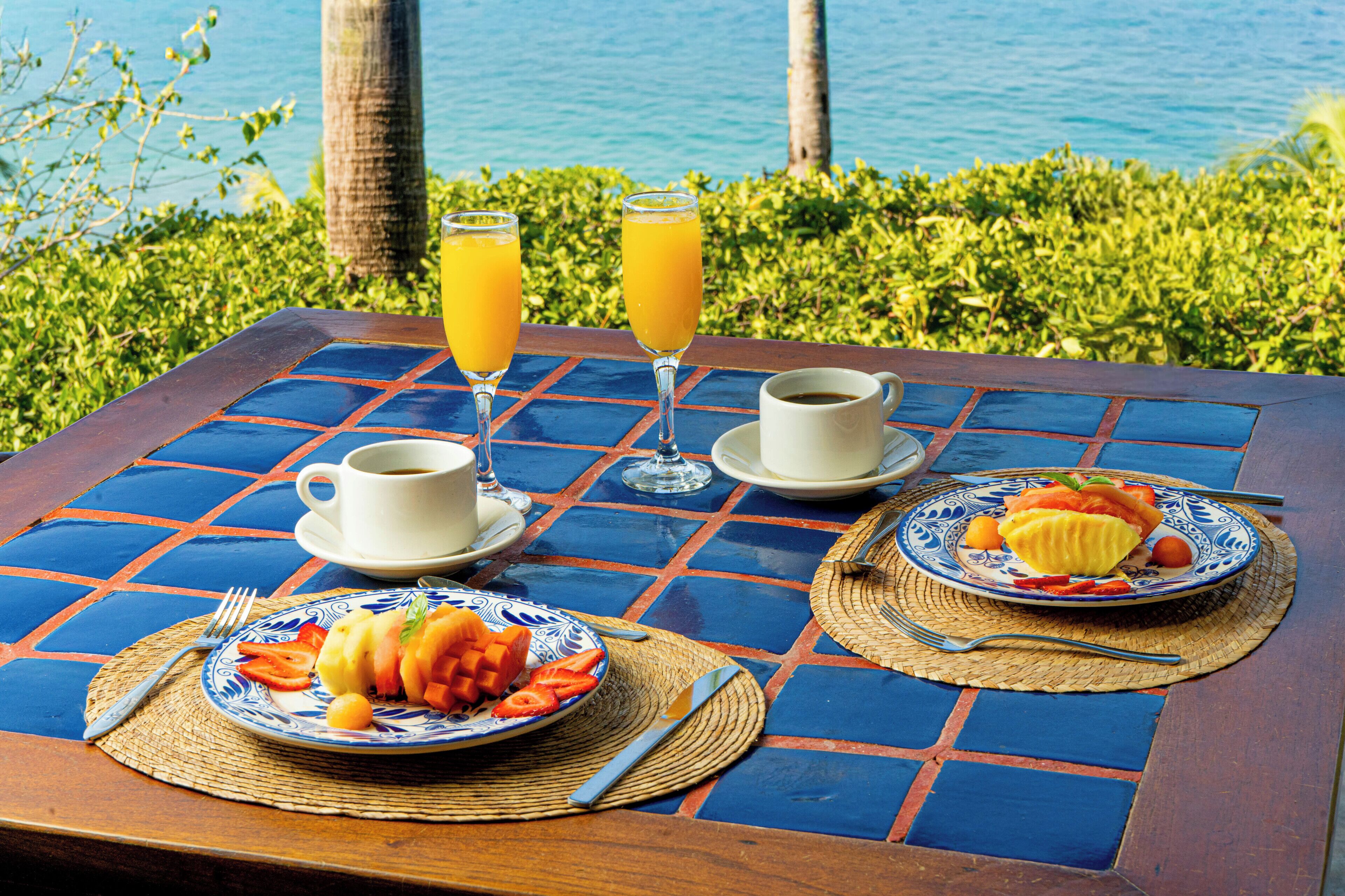 A table with a blue and orange tiled pattern, two glasses of orange juice, a cup of coffee, and plates with fruit and a dessert.