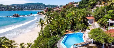 A coastal hotel with a pool, beach view, and tropical vegetation.