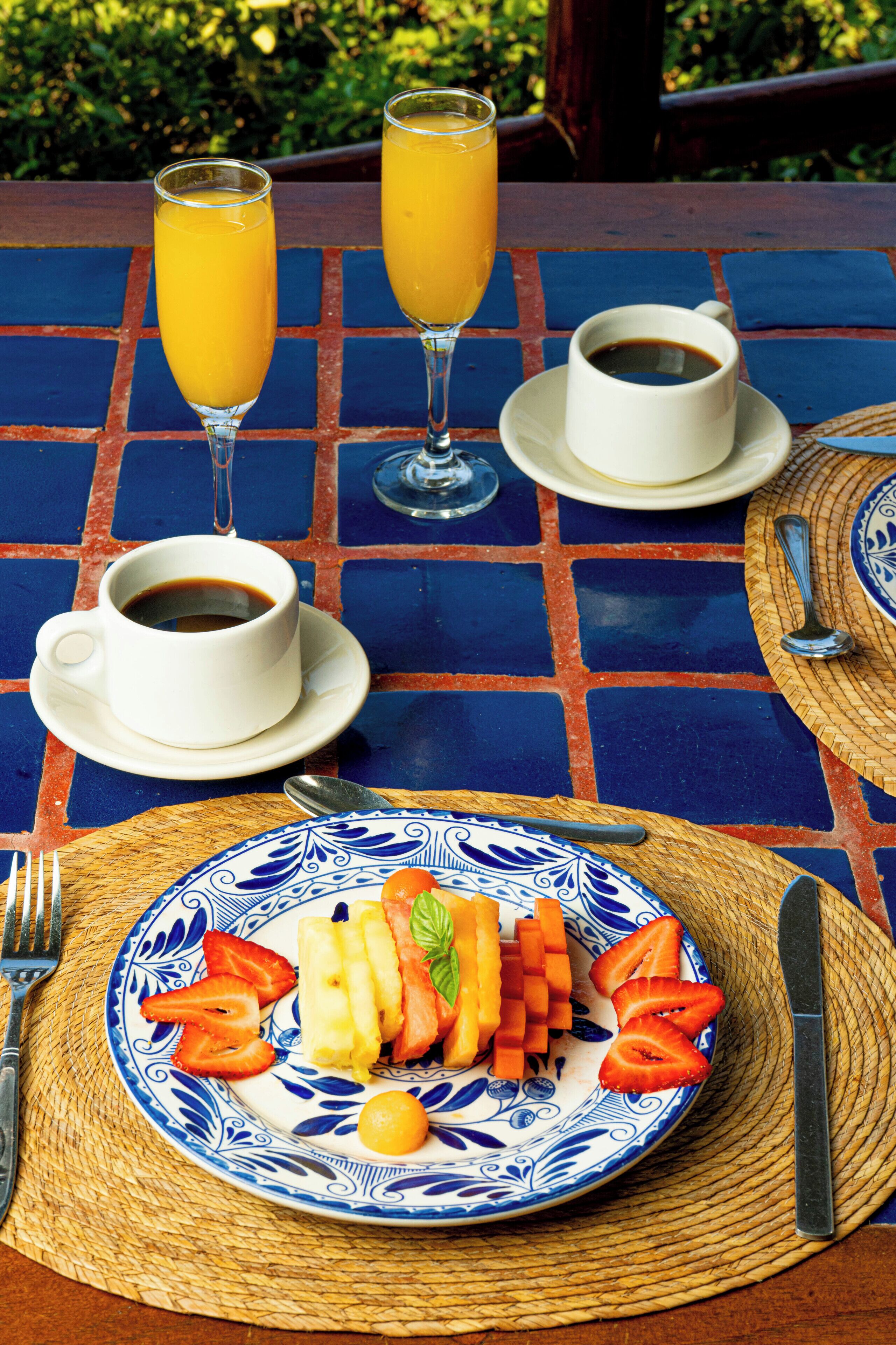 Une table dont le plateau est recouvert de carreaux bleus et oranges, une assiette de fruits, deux verres de jus d’orange, une tasse de café, ainsi qu’une fourchette et un couteau posés sur un set de table en fibres tressées.
