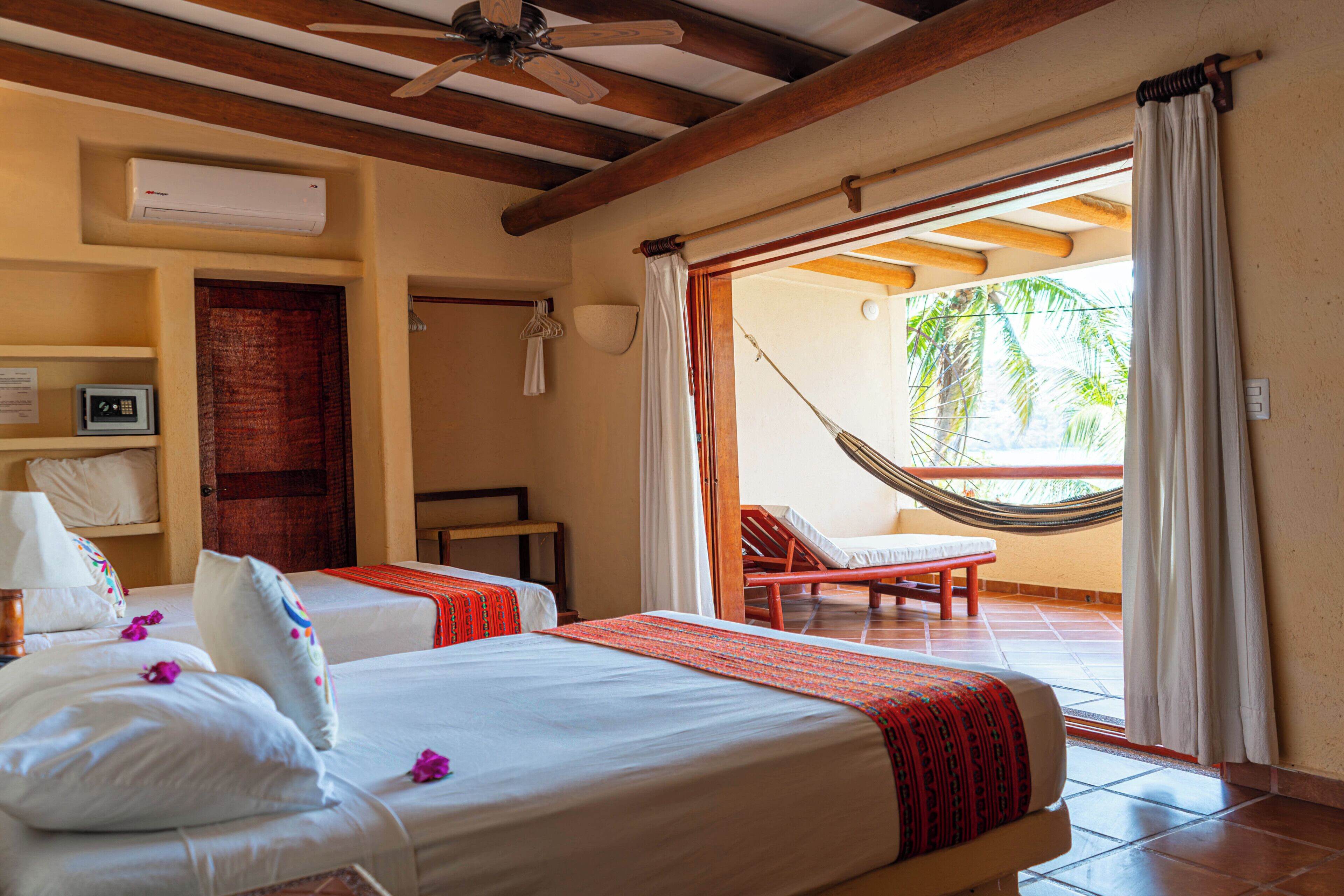 Habitación de hotel con dos camas, una hamaca, balcón con vistas a palmeras y un ventilador de techo.
