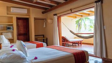 Habitación de hotel con dos camas, una hamaca, balcón con vistas a palmeras y un ventilador de techo.