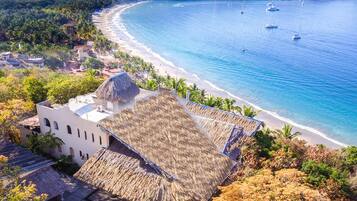 A coastal building with a thatched roof, adjacent to a beach with clear blue waters and boats.