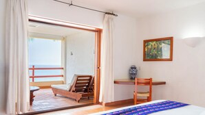 A bedroom with a bed, a desk, a chair, and a view of the sea through an open door.