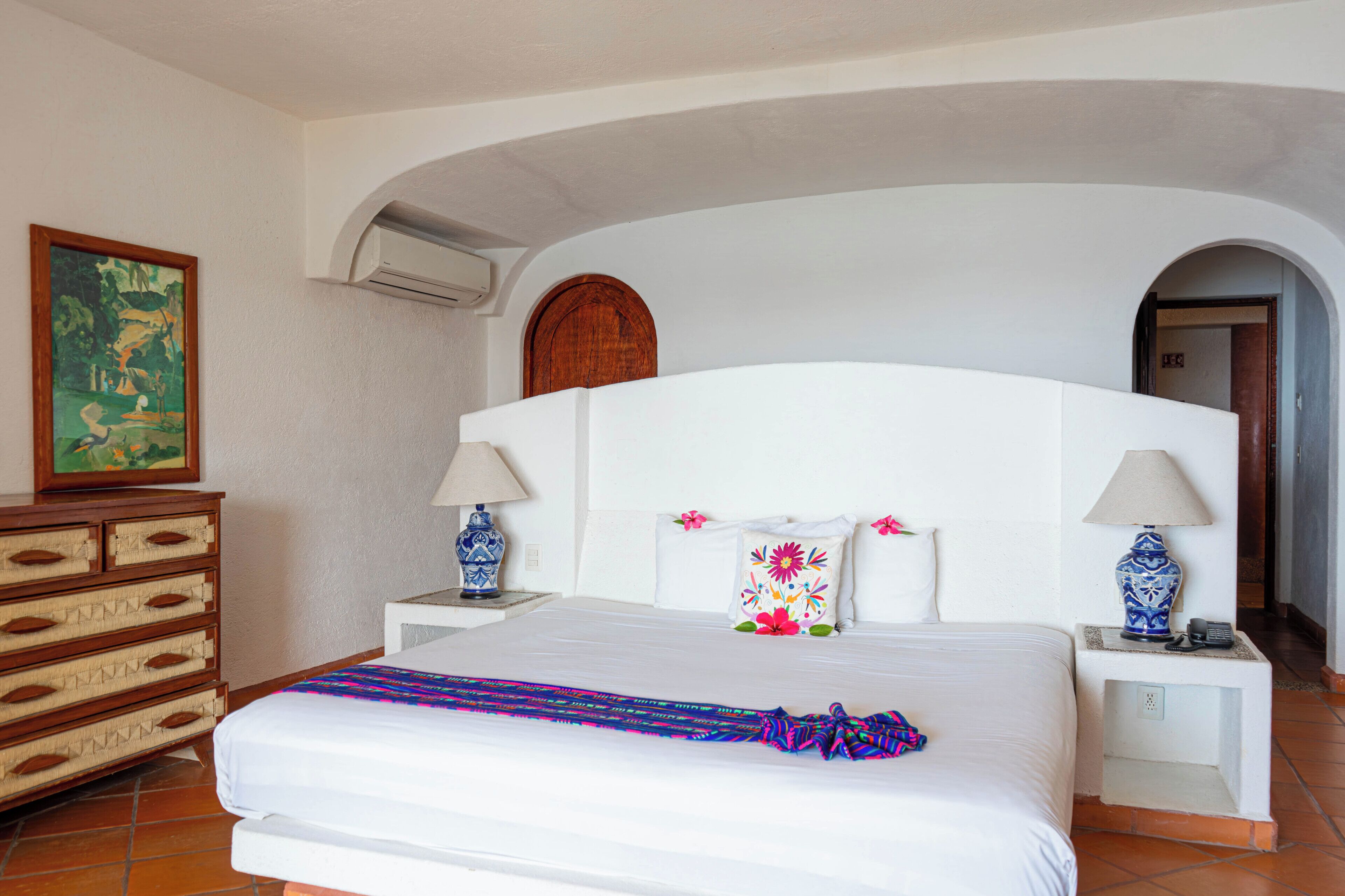Habitación de hotel con una cama grande, dos mesitas de noche con lámparas, un aparador y un cuadro en la pared, además de un arco de entrada.