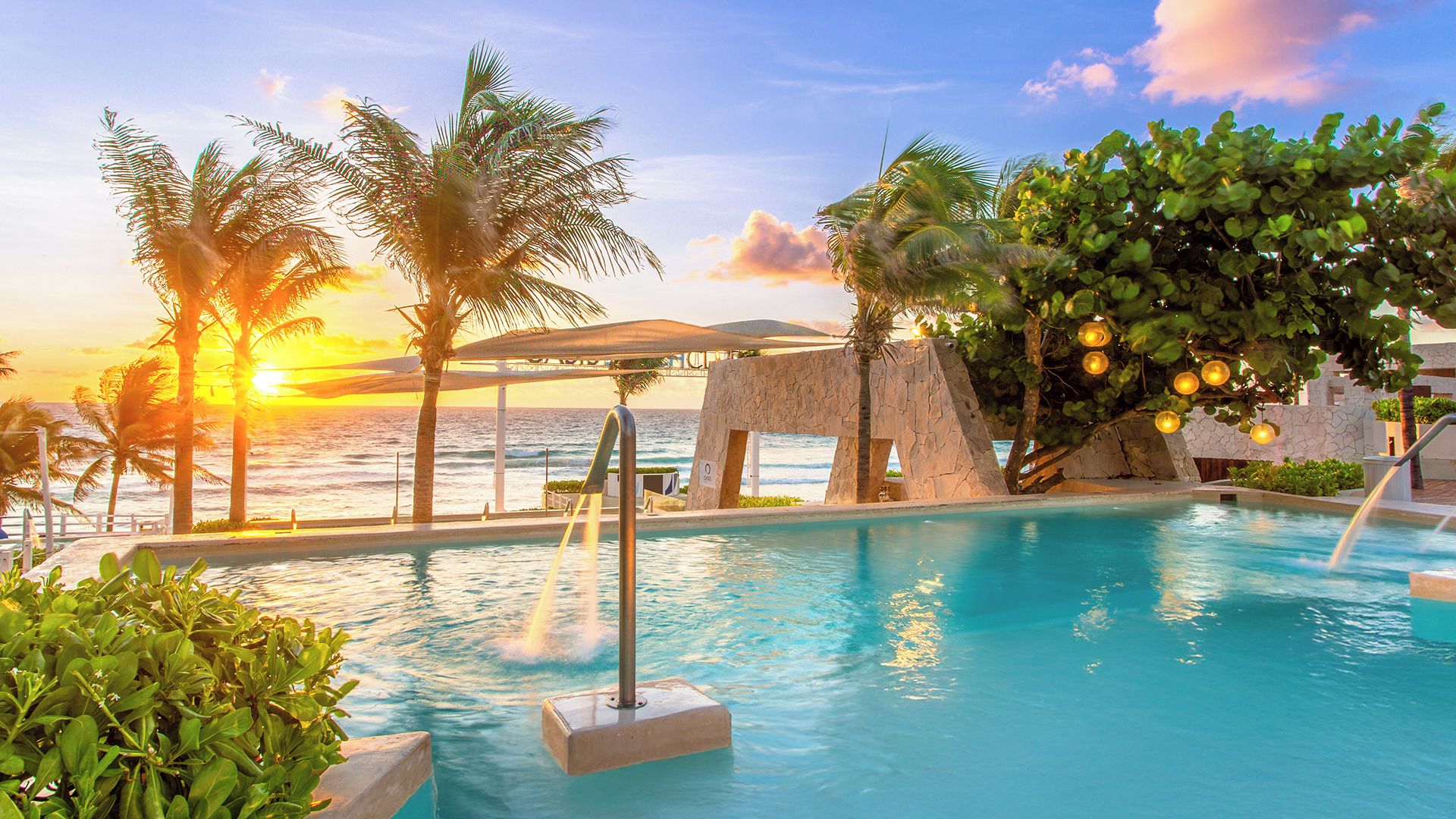 Zona de piscina con vista al atardecer, palmeras y una estructura de piedra.