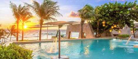 Zona de piscina con vista al atardecer, palmeras y una estructura de piedra.