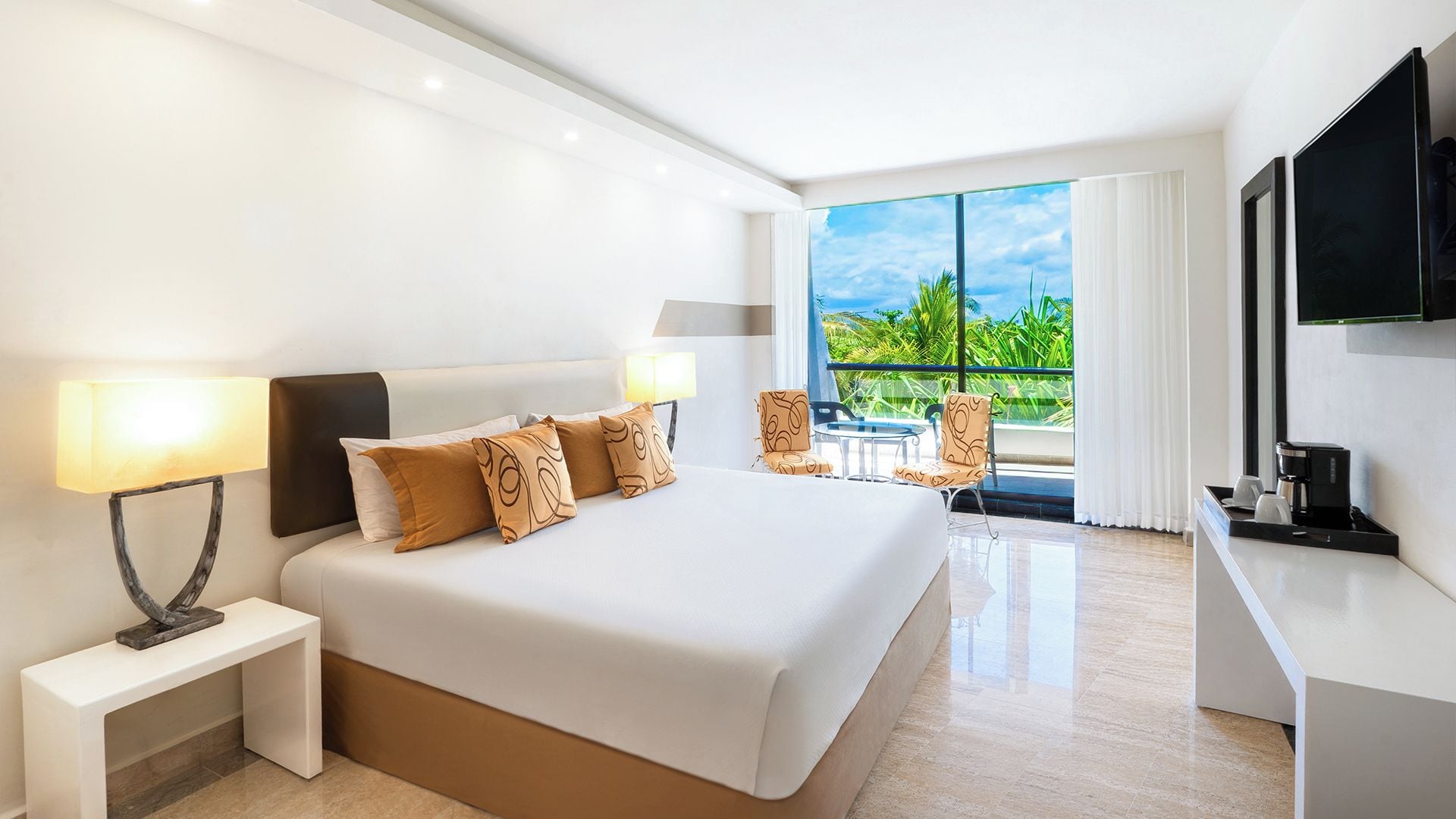 Une chambre d’hôtel avec un grand lit, un bureau avec une machine à café, une télévision et un balcon donnant sur un paysage verdoyant.