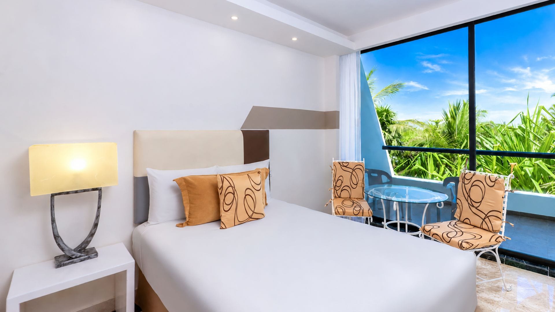Habitación de hotel con cama, mesita de noche, lámpara, silla y vistas a zonas verdes a través de amplias ventanas.