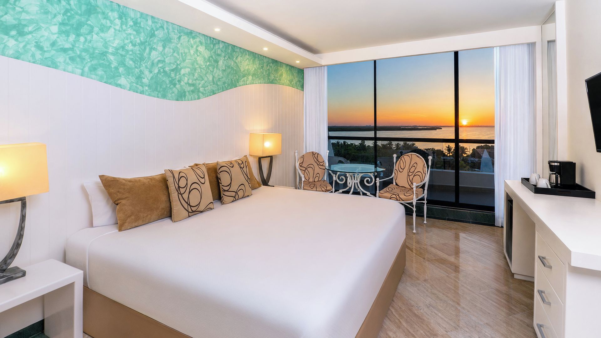 Een hotelkamer met een groot bed, een bureau, een stoel en uitzicht op de ondergaande zon door grote ramen.