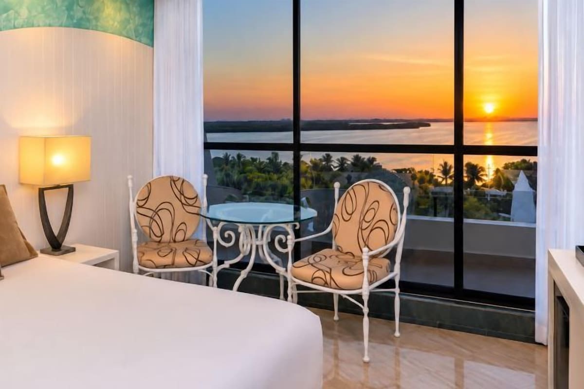 A hotel room with a bed, a table with chairs, and a large window with a sunset view.