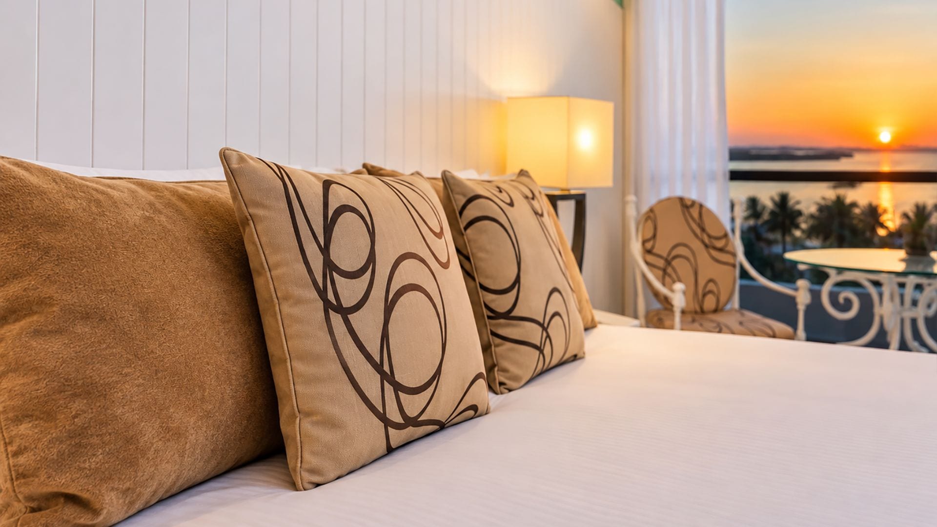 Habitación de hotel con cama, almohadas, mesita de noche, lámpara y vista al atardecer.