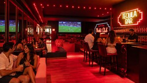 Zona de bar con un letrero de neón que dice 'TEQUILA BAR' y una pantalla grande mostrando un evento deportivo.