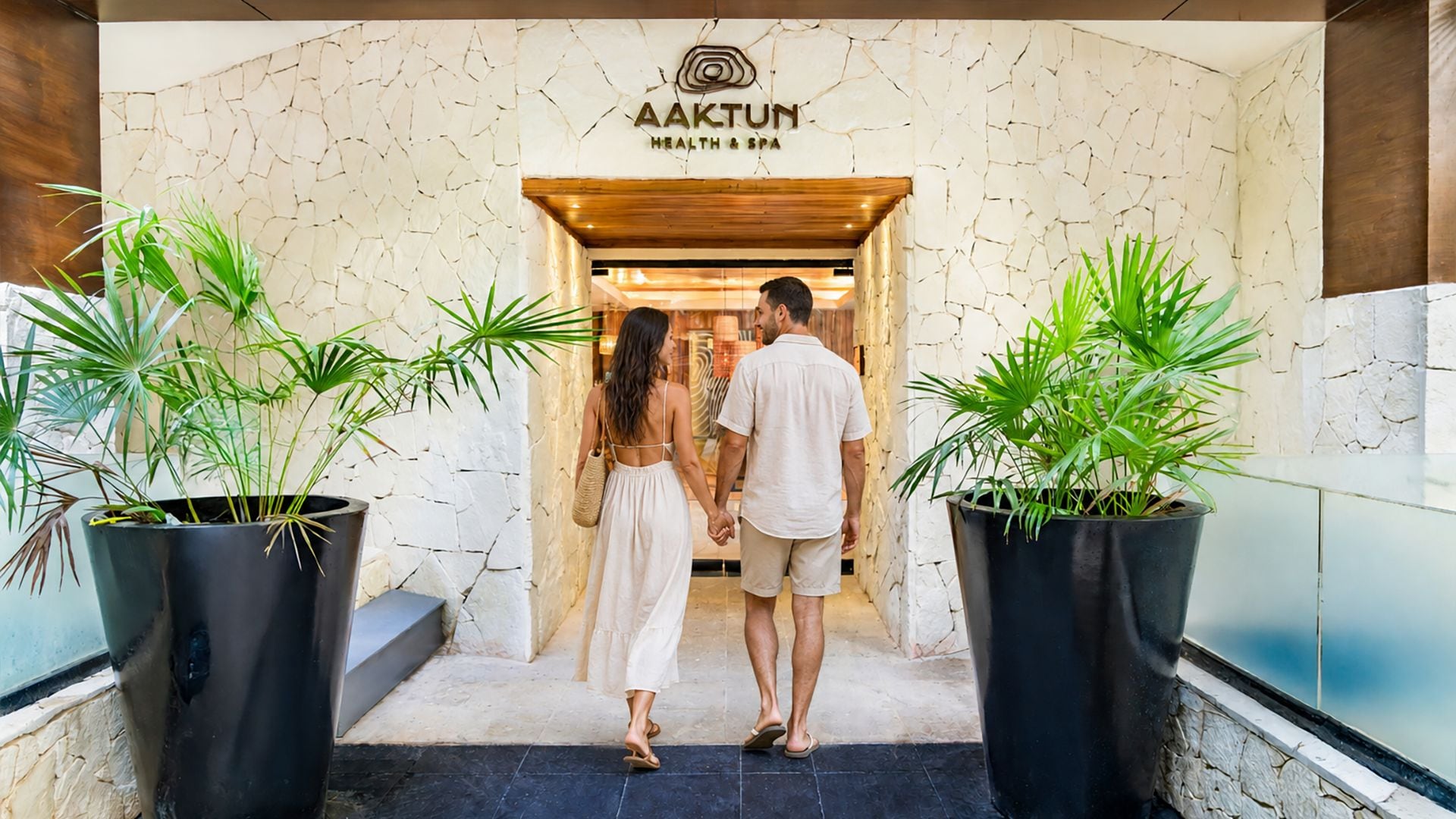 A couple walking towards a spa entrance with a cracked wall design and potted plants on either side.