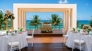 Un área de comedor al aire libre con mesas y sillas blancas, una terraza de madera y vista al océano.