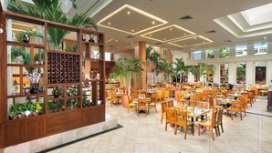 Eine großzügige Hotelhalle mit mehreren Gastronomiebereichen, einem großen Holzvitrinenregal mit verschiedenen Gegenständen und hohen Palmen.