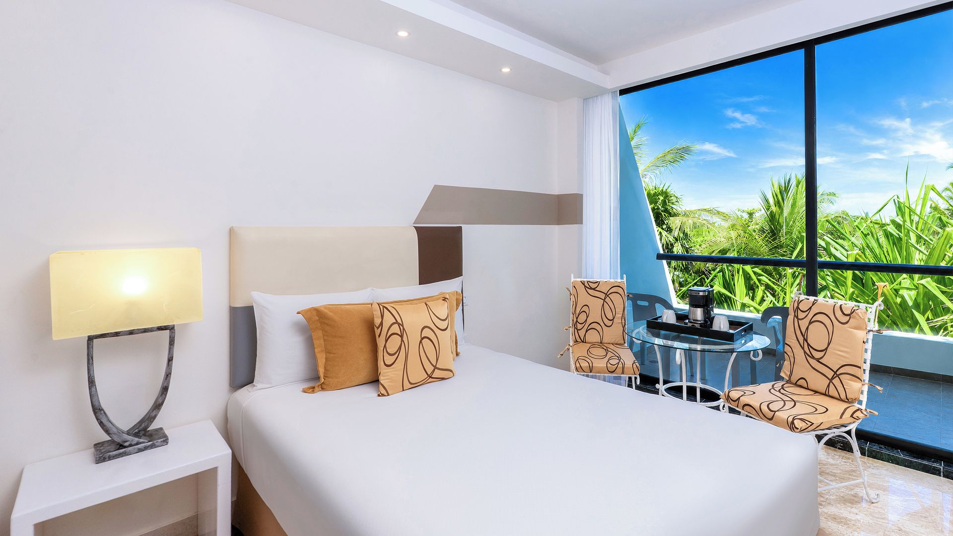 A hotel room with a bed, a nightstand, a table with chairs, and a large window with a view of greenery.
