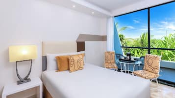 A hotel room with a bed, a nightstand, a table with chairs, and a large window with a view of greenery.