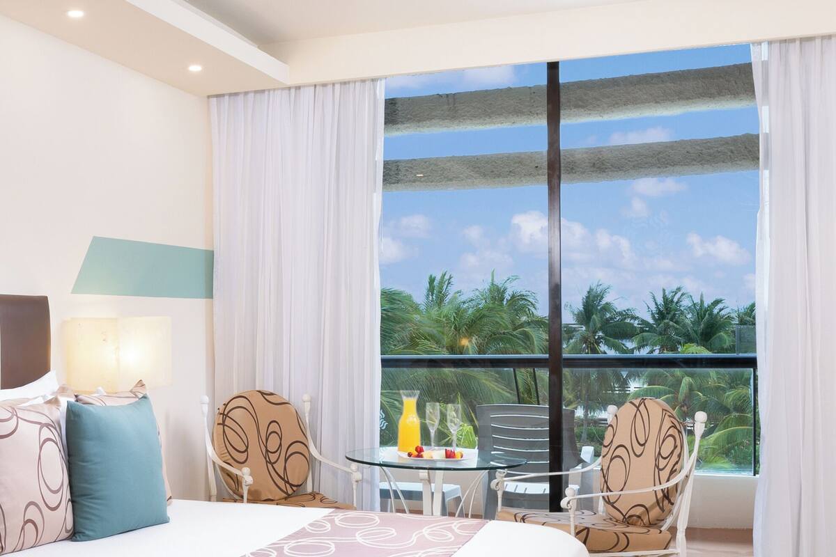 Ein Hotelzimmer mit einem großen Bett, zwei Sesseln, einem kleinen Tisch mit einem Getränk und einem Balkon mit Blick auf Palmen.
