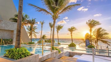 Een zwembadgedeelte van een resort met meerdere zwembaden, palmbomen en uitzicht op de oceaan bij zonsondergang.