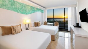 A hotel room with two beds, a desk with a TV, a balcony with a view of the sunset, and a table with chairs. - The Grand Oasis Cancun - All Inclusive (Cancun)