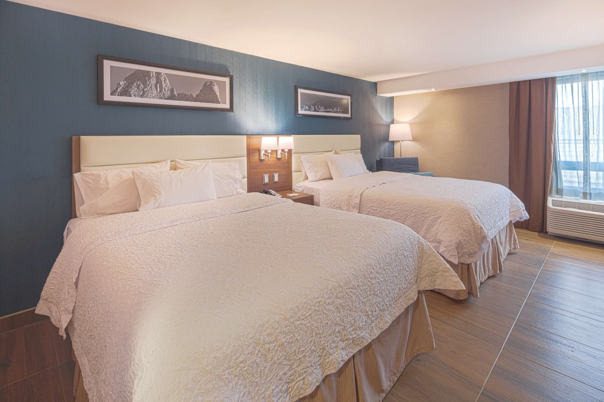 A hotel room with two beds, a nightstand with a lamp, and a window with curtains.