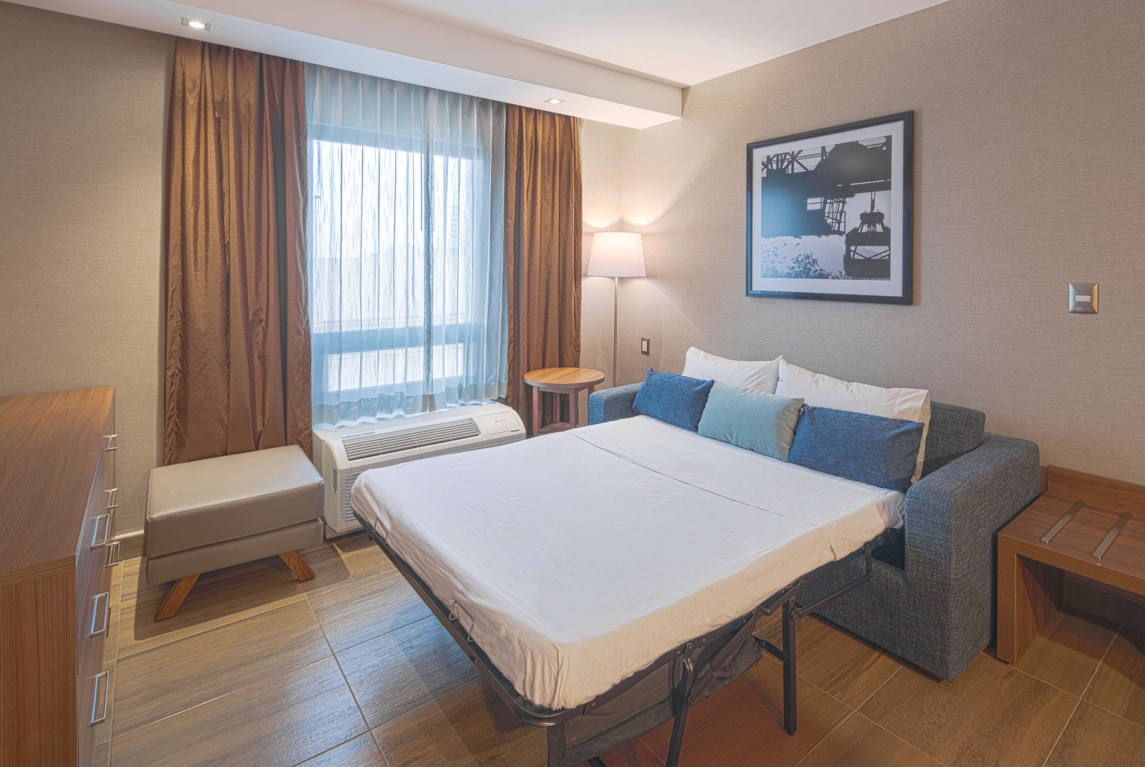 A hotel room with a bed, a bench, a desk, a lamp, and a window with curtains.