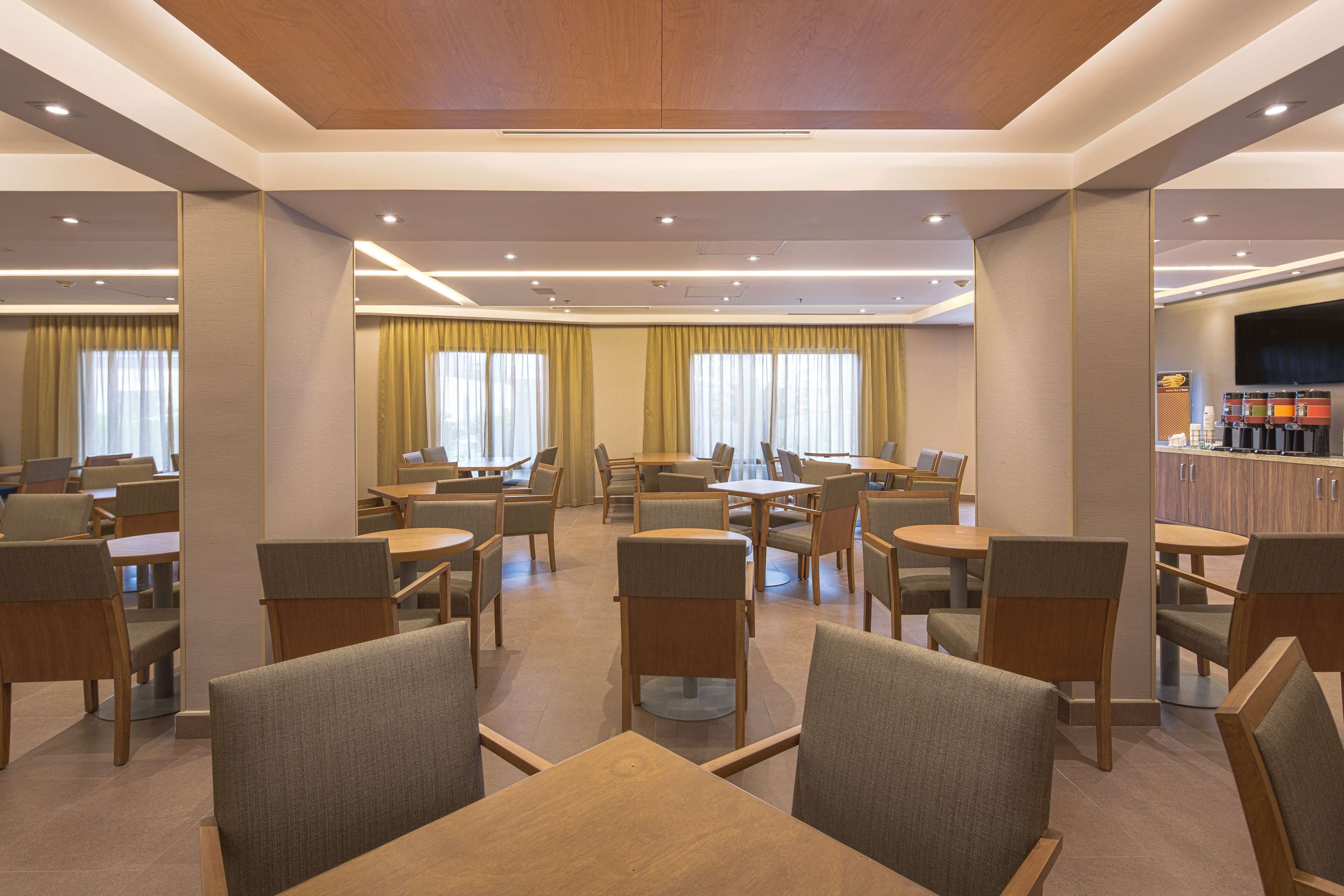 A modern hotel dining area with multiple tables and chairs, large windows with curtains, and a bar area with a flat-screen TV.