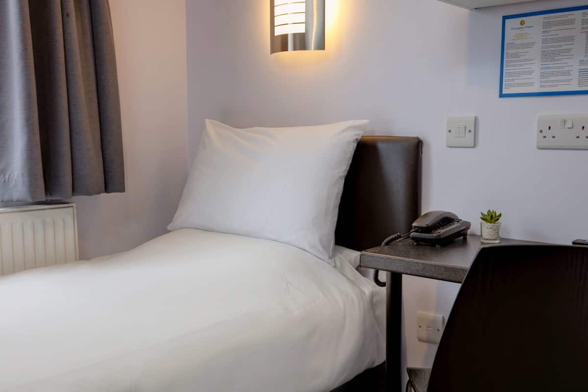Hotellihuone, jossa on sänky, kirjoituspöytä, jossa on puhelin, pieni kasvi ja seinälle asennettu valaisin.
