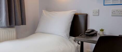 A hotel room with a bed, a desk with a telephone, a small plant, and a wall-mounted light fixture.