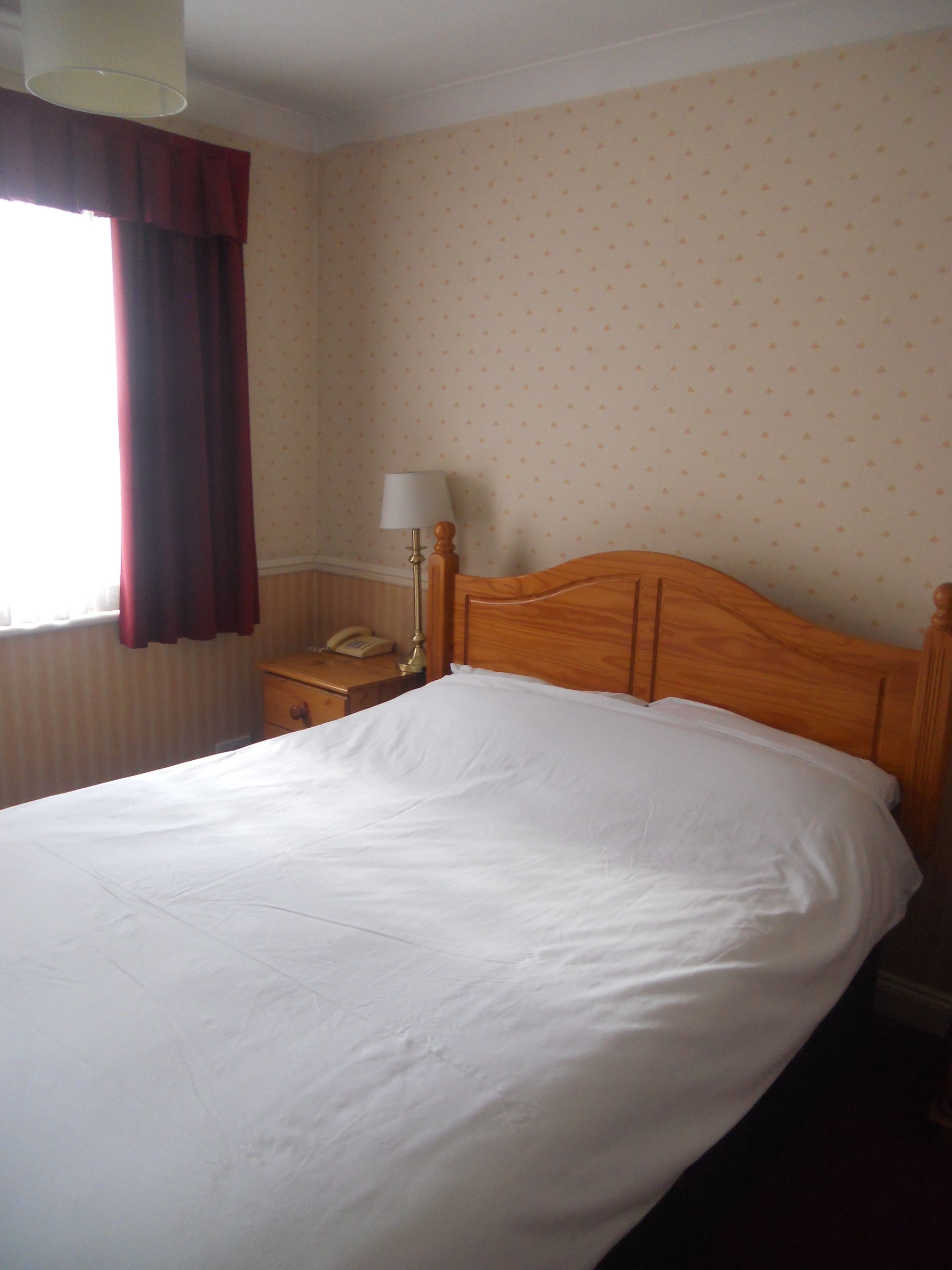 A neatly made bed in a hotel room with a wooden headboard, a bedside table with a lamp, and a patterned wallpaper.