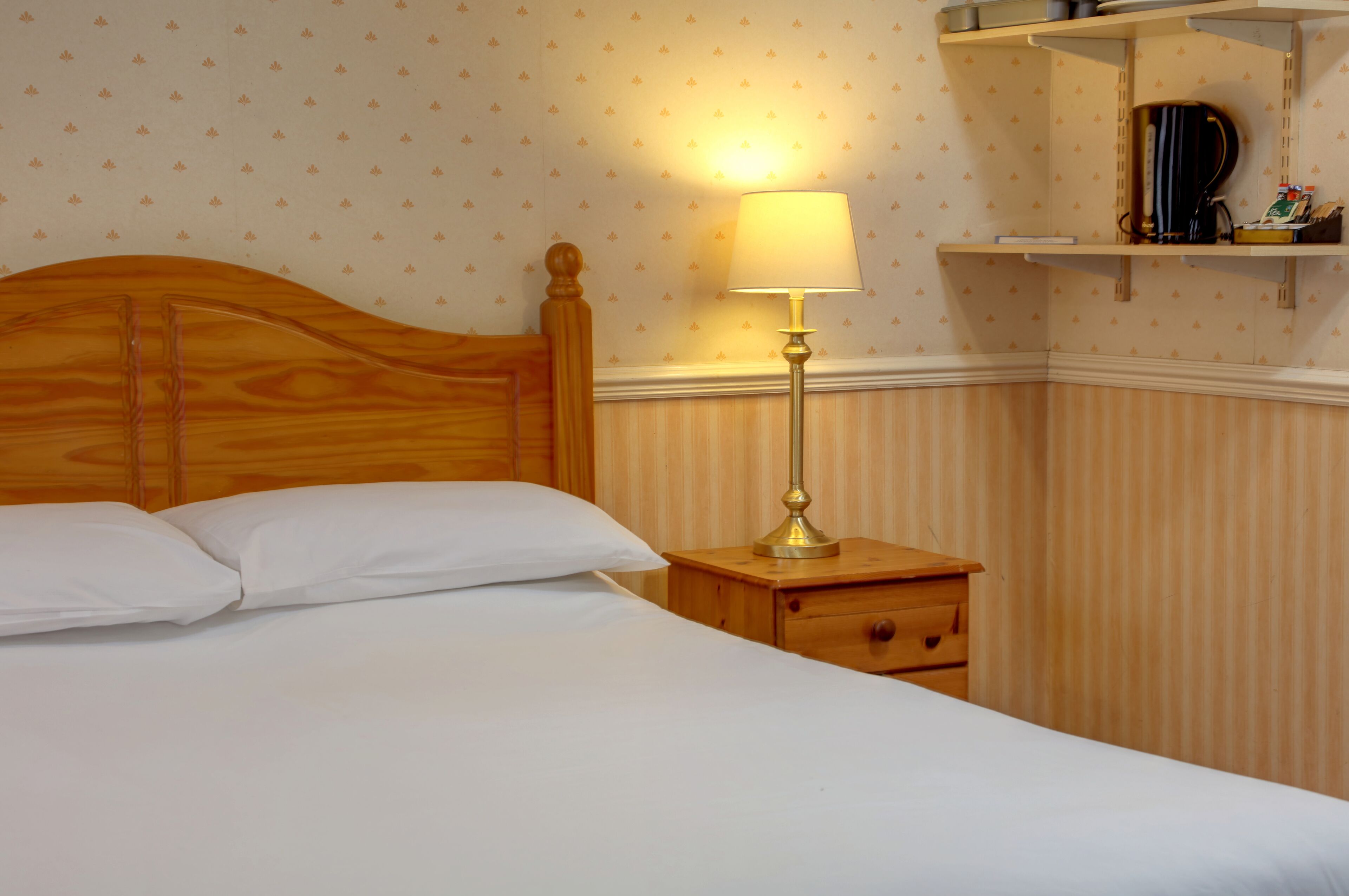 A neatly made bed with white linens, a wooden headboard, a bedside table with a lamp, and a shelf with items.