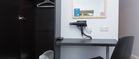 A hotel room with a dark wooden door, a small desk with a hairdryer, a mirror, and a wall-mounted fan.