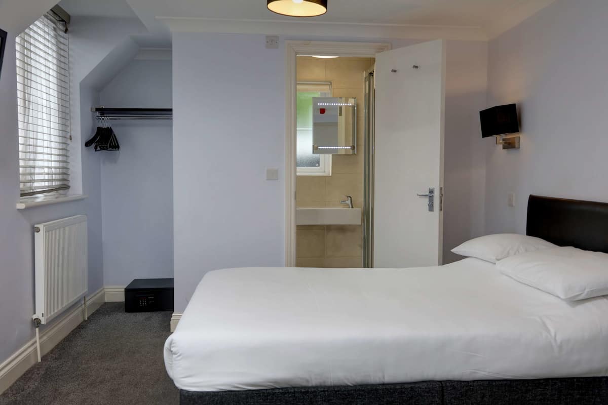 Hotellihuone, jossa on suuri sänky, yöpöytä, peili, patteri, ikkuna, jossa on sälekaihtimet, ja seinälle asennettu televisio.