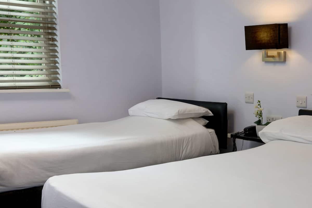 Hotellihuone, jossa on kaksi erillistä sänkyä, ikkuna, jossa on sälekaihtimet, yöpöytä lampun kanssa ja seinään kiinnitetty valaisin.