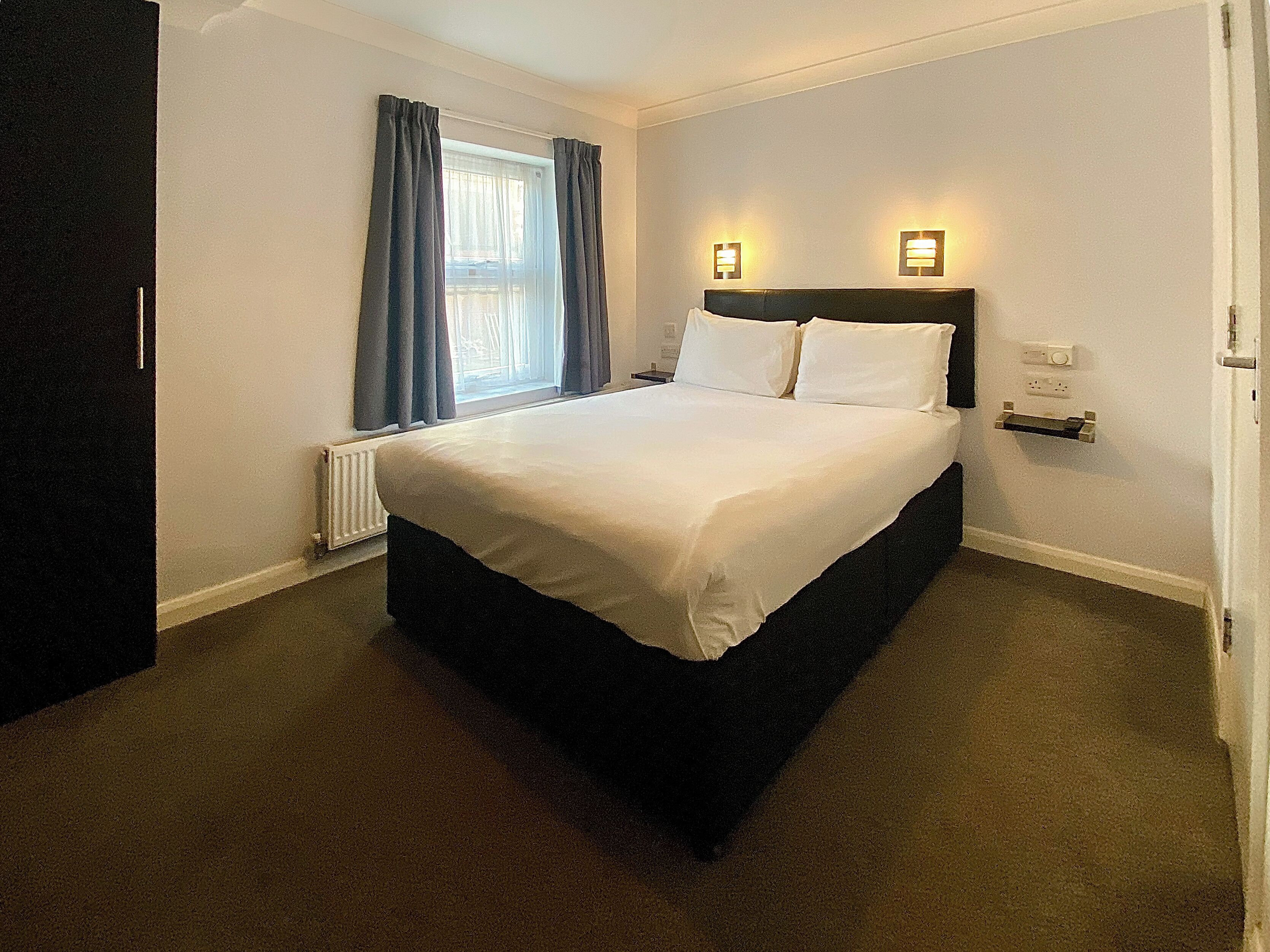 A hotel room with a large bed, a window with curtains, a bedside table with a lamp, and a thermostat on the wall.