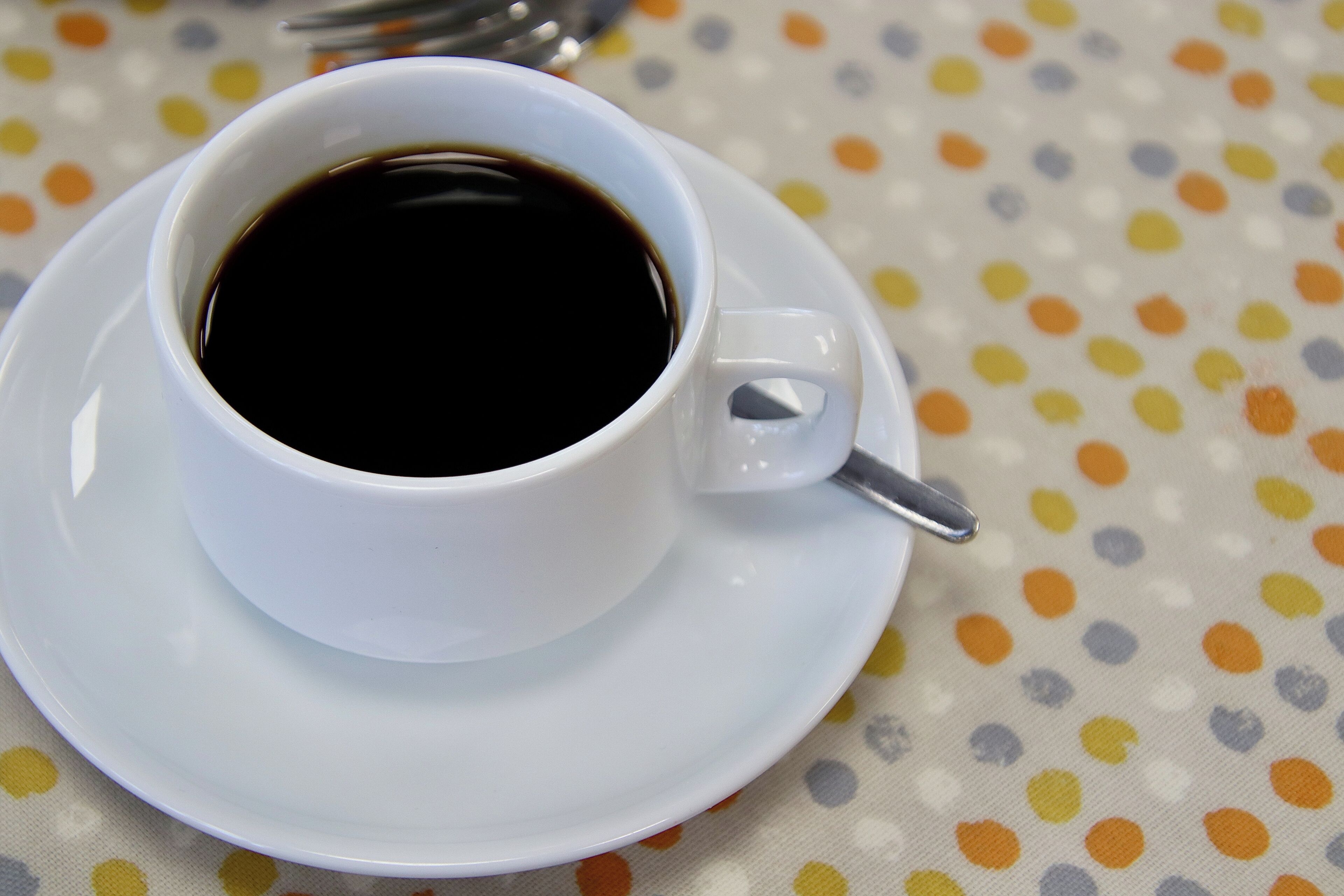 Una taza blanca de café sobre un plato, con una cuchara, colocada en una mesa con mantel de lunares.