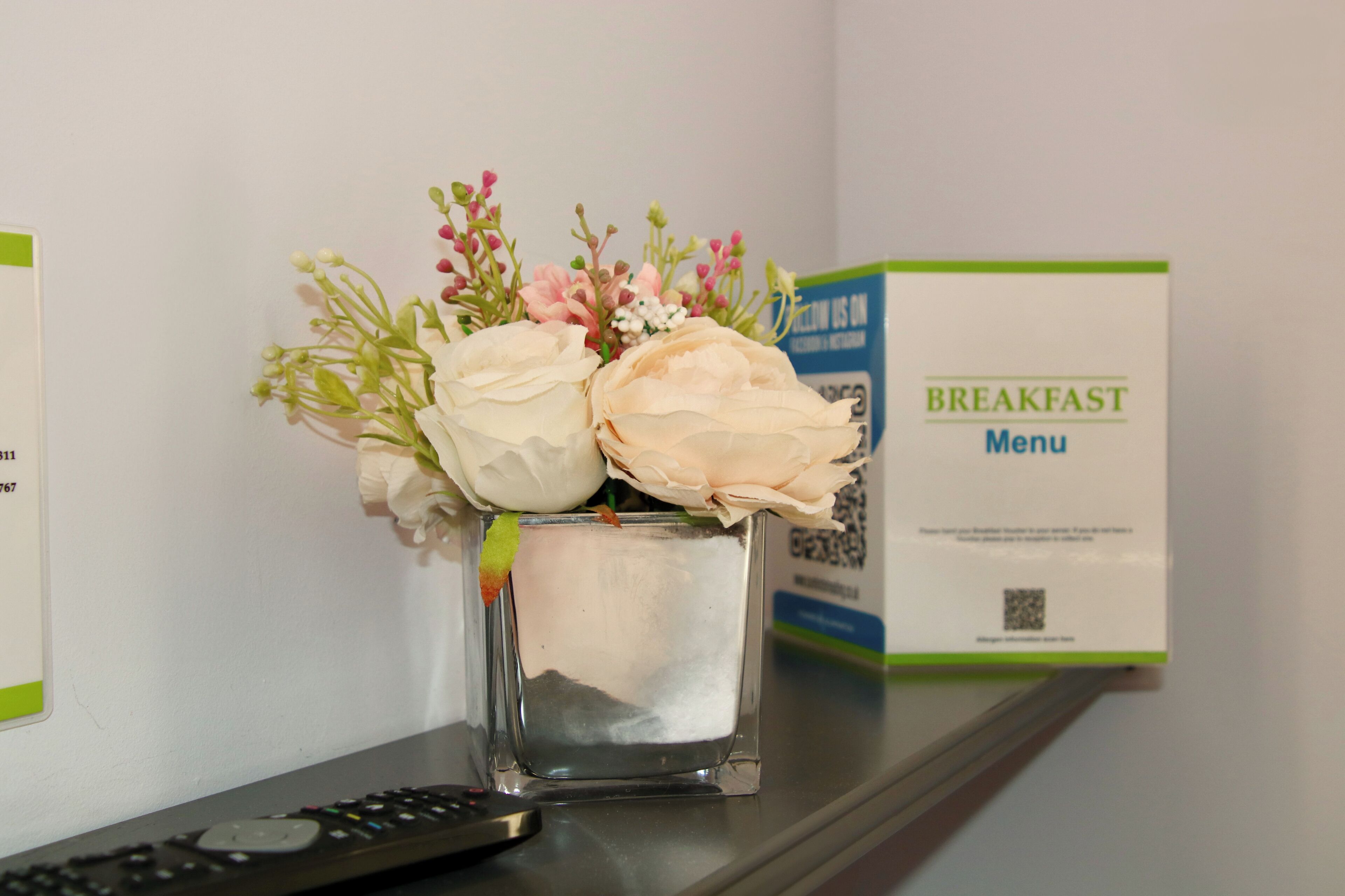 Ein kleiner, quadratischer Blumenvasen mit künstlichen Blumen auf einem Regal, einer Fernbedienung und einem Frühstücksmenü-Schild.