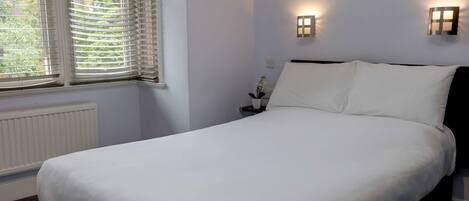 A neatly made bed in a room with white walls, two bedside lamps, and a window with blinds.
