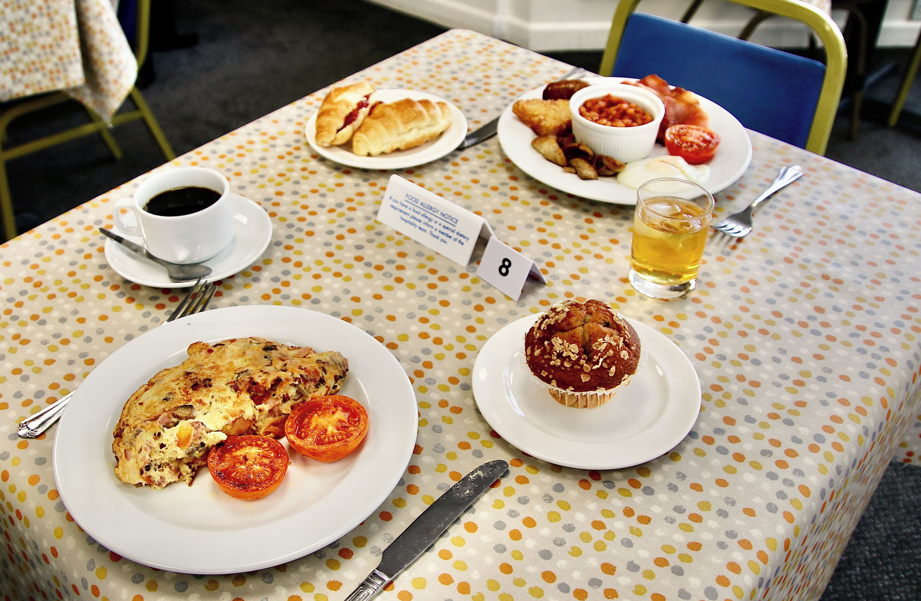 A table with a polka dot tablecloth, plates of food, a cup of coffee, a glass of tea, and a sign indicating a food allergy notice.