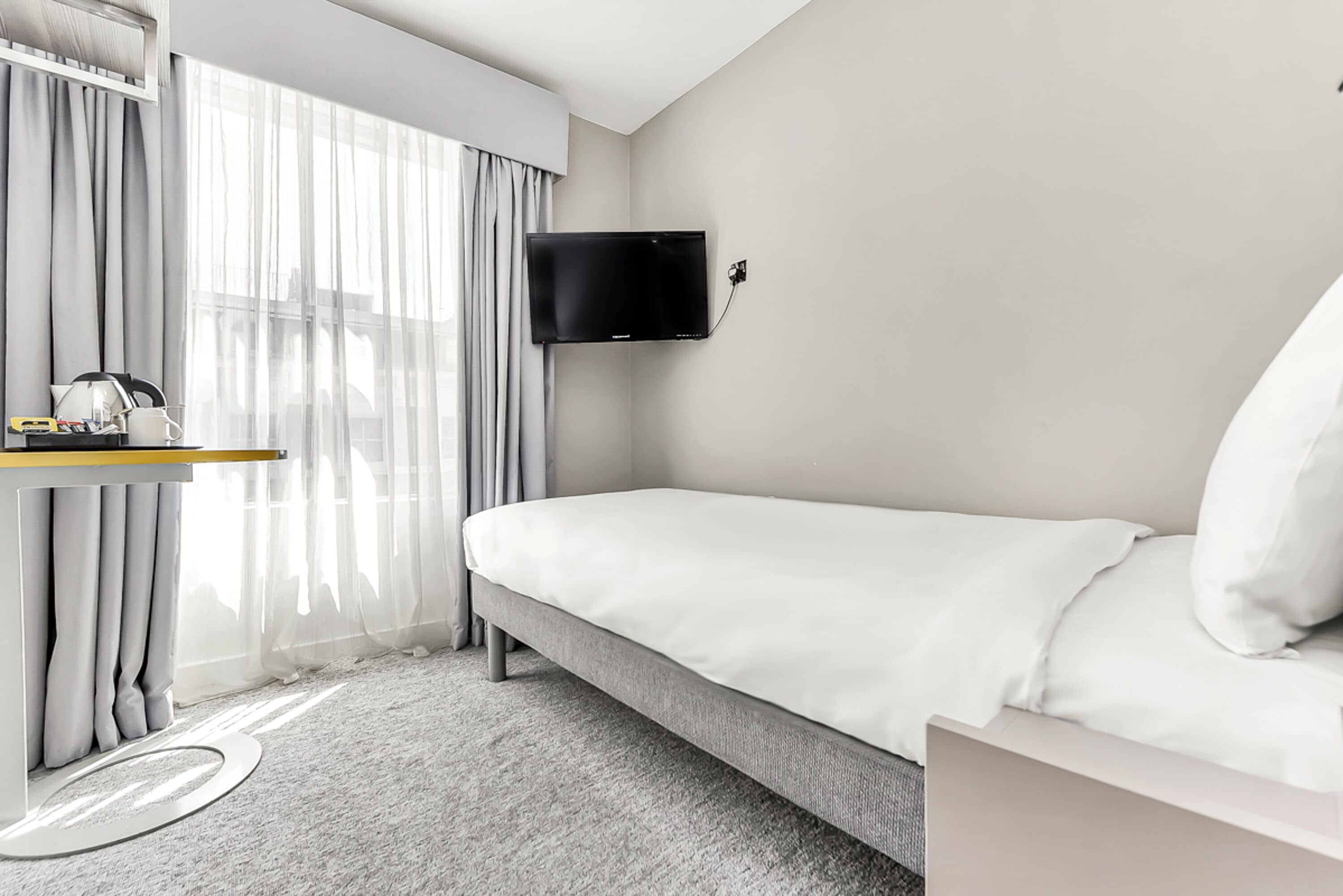 A modern hotel room with a single bed, a mounted TV, a small desk with a lamp, and a window with sheer curtains.