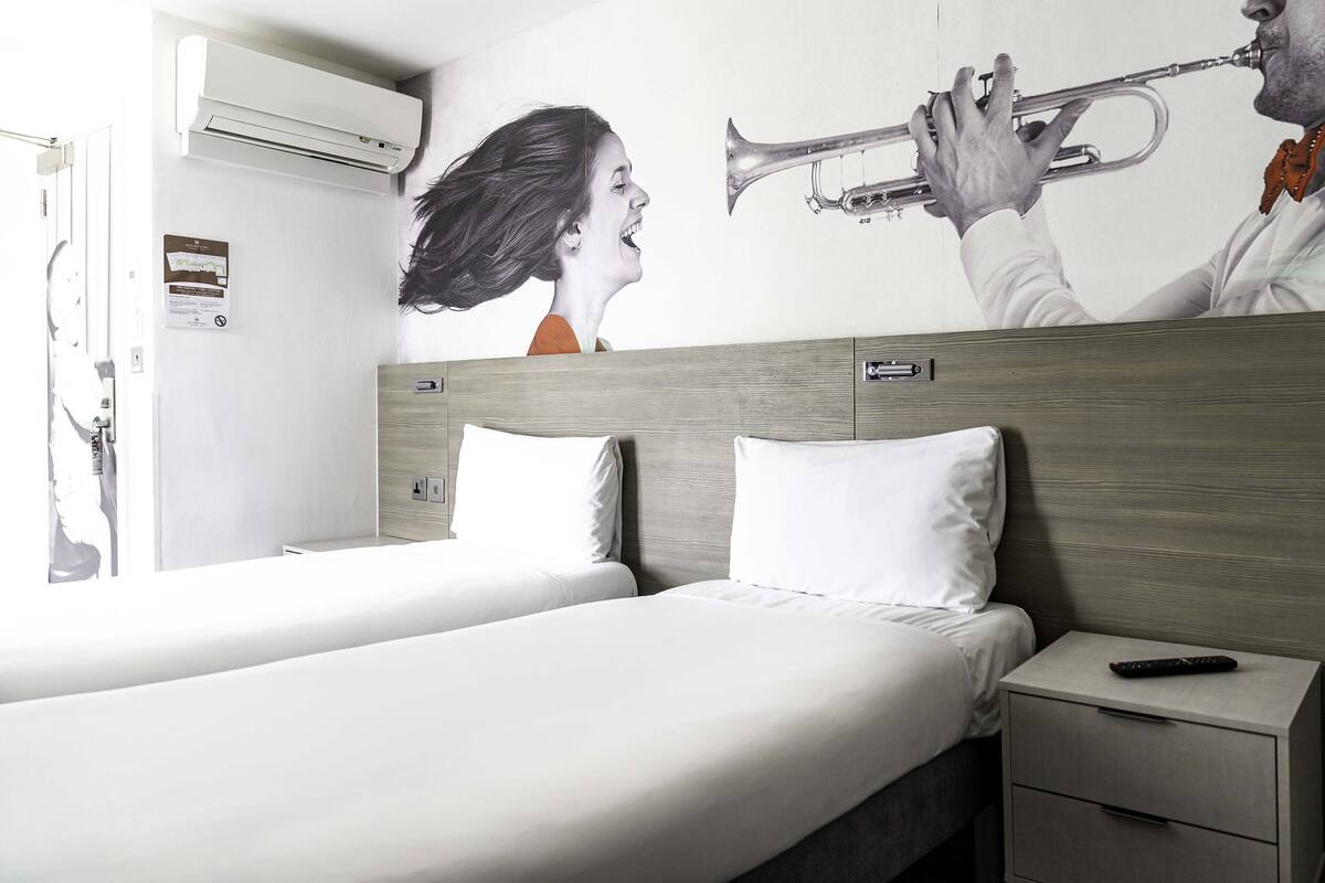 Een hotelkamer met een bed, een nachtkastje, een afstandsbediening en een muurschildering van een persoon die trompet speelt.