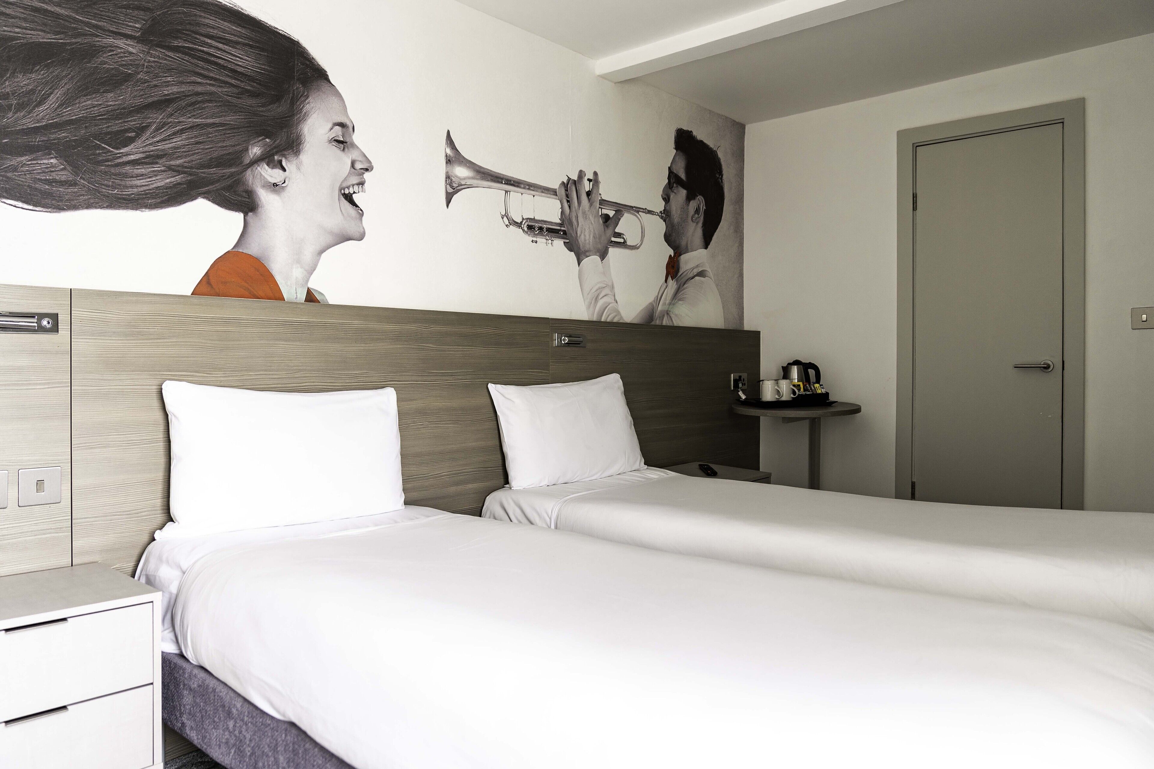 Habitación de hotel con dos camas, cabecera de madera, mesita de noche pequeña con lámpara y un cuadro en la pared que muestra a una persona tocando una trompeta.